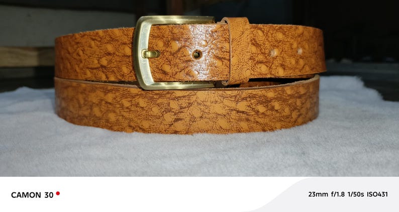 May include: A textured, tan leather belt with a gold-colored buckle. The belt is rolled up and resting on a white surface. The belt has a detailed pattern and a classic design.