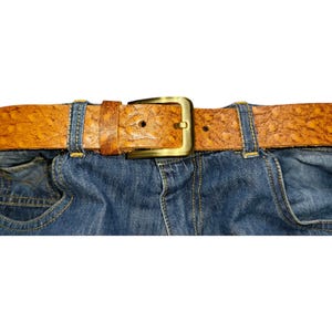 May include: A close-up of blue denim jeans with a brown leather belt. The belt has a textured surface and a gold-colored buckle. The belt is secured through the jean's belt loops.