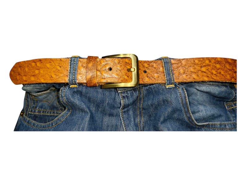 May include: A close-up of blue denim jeans with a brown leather belt. The belt has a textured surface and a gold-colored buckle. The belt is secured through the jean's belt loops.