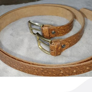 May include: Two brown leather belts with textured surfaces and brass-colored buckles. The belts are arranged in a circular pattern, showcasing their design and craftsmanship. The image is well-lit, highlighting the details of the belts.