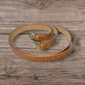 May include: Two brown leather belts with textured patterns and brass-colored buckles are displayed on a wooden surface. The belts are coiled and arranged to showcase their design and hardware.