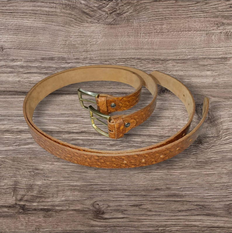 May include: Two brown leather belts with textured patterns and brass-colored buckles are displayed on a wooden surface. The belts are coiled and arranged to showcase their design and hardware.