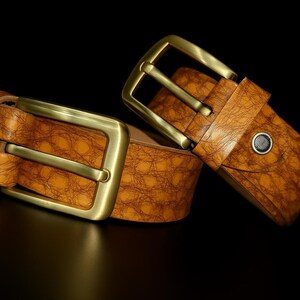 May include: Two brown leather belts with gold-colored buckles are displayed against a black background. The belts have a textured surface, and the buckles are rectangular. The belts are likely for pants or trousers.