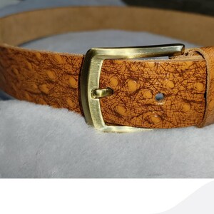 May include: A brown leather belt with a textured pattern and a gold-colored rectangular buckle. The belt is displayed on a soft, white surface, showcasing its details and craftsmanship. The belt is a fashion accessory.