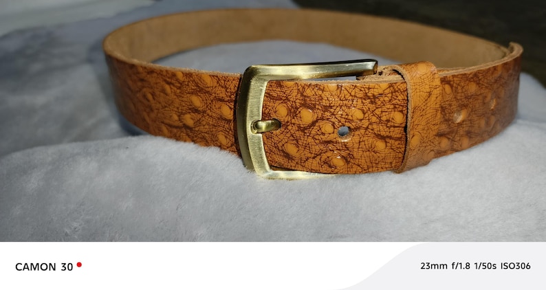 May include: A brown leather belt with a textured pattern and a gold-colored rectangular buckle. The belt is displayed on a soft, white surface, showcasing its details and craftsmanship. The belt is a fashion accessory.