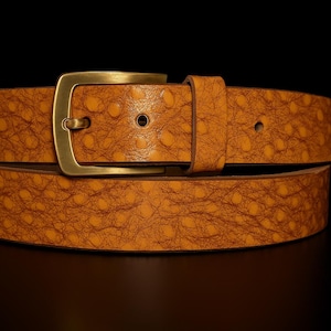 May include: A tan leather belt with a textured pattern and a brass buckle. The belt is made of a single piece of leather and has a classic design. The buckle is rectangular and has a simple, elegant look. The belt is suitable for casual or formal wear.
