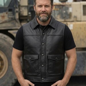 May include: A black leather vest with a collared neckline and two front pockets. The vest has a button-up closure and is worn over a black t-shirt. The person is standing outdoors with a construction vehicle in the background.