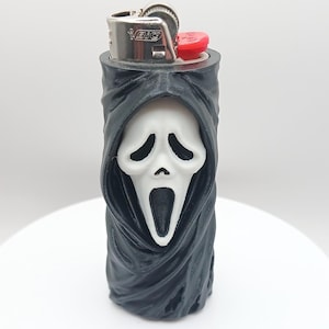 May include: A black Bic lighter encased in a three-dimensional sleeve featuring a white Ghostface mask with a black hooded robe. The metal ignition mechanism is visible at the top, with the Bic logo clearly printed on the silver metal surface.