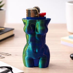 May include: A blue and green decorative lighter holder in the shape of a female torso. The holder has a blue base and holds a gold and red lighter. The holder is on a wooden desk.
