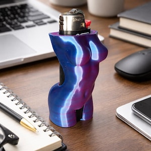 May include: A decorative lighter case in the shape of a female torso. The case is a gradient of blue and purple. The lighter is silver with a red button. The item is on a wooden desk with office supplies.
