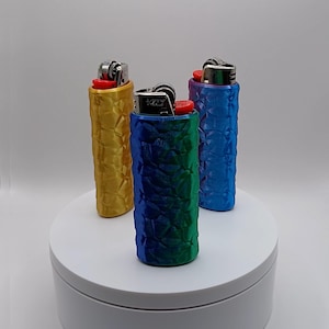 May include: Three lighters in textured cases, in gold, blue-green gradient, and blue. The lighters are on a white, circular platform against a white backdrop. The cases have a geometric pattern.
