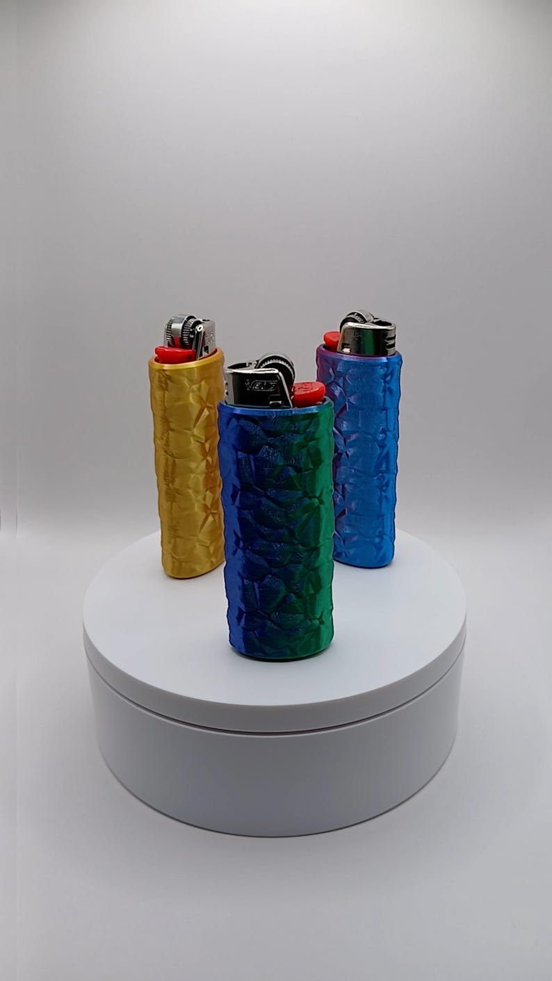 May include: Three lighters in textured cases, in gold, blue-green gradient, and blue. The lighters are on a white, circular platform against a white backdrop. The cases have a geometric pattern.