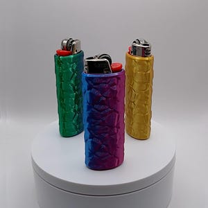 May include: Three lighters in textured cases, each with a different color. The cases are green, blue-purple gradient, and gold. The lighters are on a white platform against a white background.