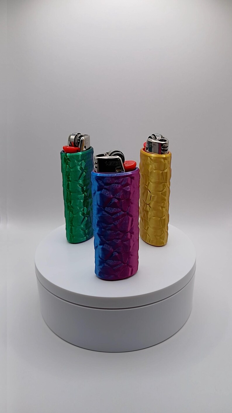 May include: Three lighters in textured cases, each with a different color. The cases are green, blue-purple gradient, and gold. The lighters are on a white platform against a white background.