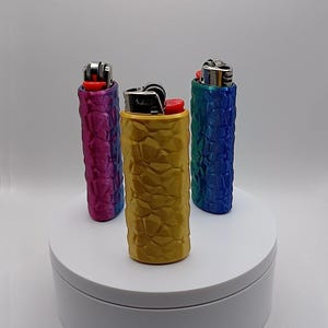 May include: Three lighters in textured cases, displayed on a white platform. The cases are in shades of gold, blue, and purple. The lighters have red ignition buttons and silver metal components.