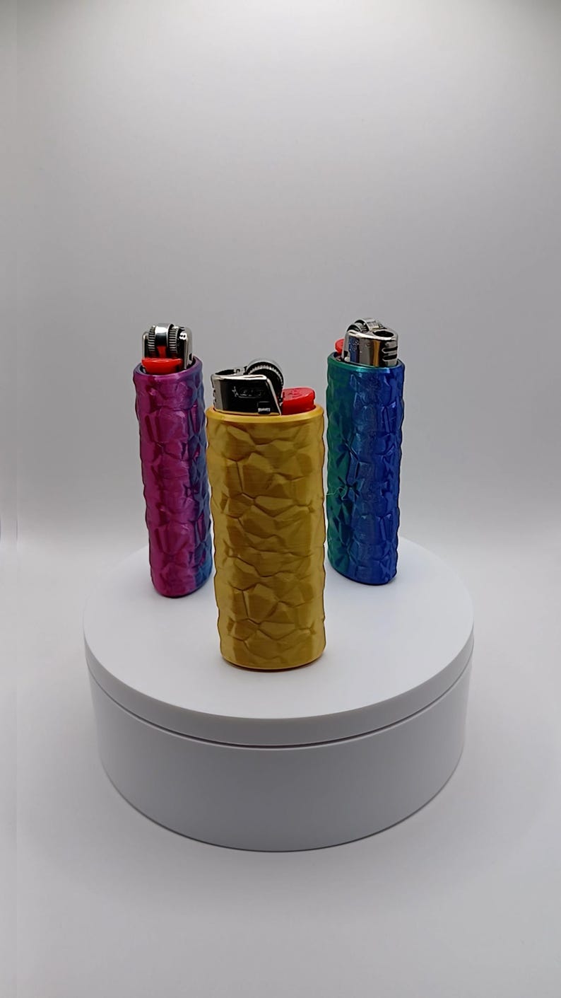 May include: Three lighters in textured cases, displayed on a white platform. The cases are in shades of gold, blue, and purple. The lighters have red ignition buttons and silver metal components.