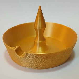 May include: A gold-colored ashtray with a textured rim and a central conical spike. The ashtray has a circular design with a small notch on the side. The ashtray is made of a solid material.