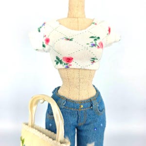 May include: A miniature mannequin torso wearing a white short-sleeved crop top with a green diamond pattern and pink floral print, paired with blue denim jeans featuring small blue rhinestones and a cream-colored handbag with floral embroidery.