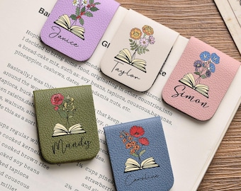 May include: A collection of five leather book page holders in various colors: lavender, cream, pink, olive green, and blue. Each features a floral design above an open book illustration and a name in cursive.