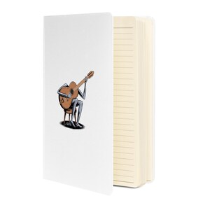 Könnte beinhalten: Weißes Notizbuch mit einer Cartoon-Illustration einer Figur, die Gitarre spielt. Die Figur sitzt auf einem Stuhl, mit der Gitarre in den Armen. Das Notizbuch hat linierte Seiten.