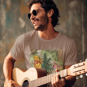 May include: A light brown t-shirt featuring a cartoon image of Winnie the Pooh and Piglet smoking marijuana. The image shows Winnie the Pooh smoking a joint next to a marijuana plant. The man in the image is wearing sunglasses and playing a guitar.