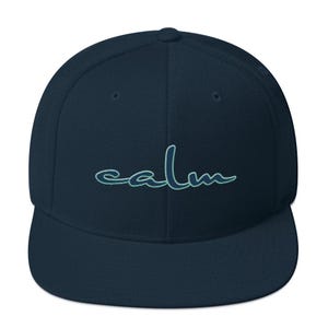 May include: A navy blue flat-brimmed baseball cap with the word "calm" embroidered in a teal script across the front. The cap has a structured fit and a rounded crown.