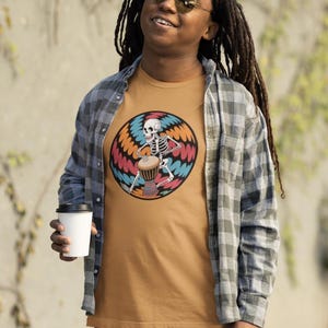 Può includere: T-shirt color senape con uno scheletro che suona un tamburo djembe all'interno di un design circolare colorato. La persona indossa una camicia a quadri e occhiali da sole, tenendo in mano una tazza di caffè bianca. Il tema generale è la musica e l'arte.