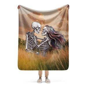 May include: A throw blanket featuring a skeletal couple embracing in a field of tall grass. The skeletons are depicted in a romantic pose, with the female skeleton's hair flowing in the wind. The background is a soft, muted orange and brown.