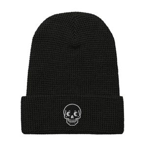 May include: Black beanie with a waffle knit texture. The beanie features a white embroidered skull design on the front. The skull has detailed eye sockets and teeth. The beanie is a winter accessory.