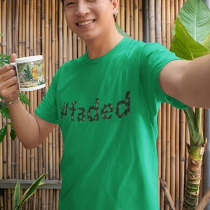 May include: A person wearing a green t-shirt with the word "#faded" in a dark green, leafy font. The person is holding a white mug with a colorful cartoon design. The background features bamboo and plants.