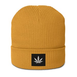 May include: Mustard yellow beanie with a ribbed knit texture. A black square patch on the front features a white embroidered cannabis leaf design. The beanie is a casual accessory.