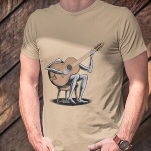 May include: A tan t-shirt with a graphic of a skeleton playing a guitar. The skeleton is sitting on a stool and holding a guitar with its bony fingers.