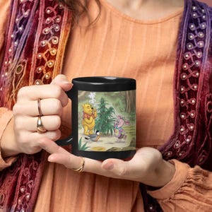 May include: Black ceramic mug featuring a whimsical illustration of a yellow bear, a pink pig, and a bird in a forest setting. The bear is smoking a plant, and the pig is wearing a tie-dye outfit. The mug has a black handle.