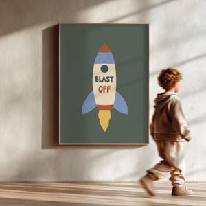 May include: A framed print featuring a retro-style rocket ship illustration. The rocket is primarily cream-colored with blue, red, and yellow accents. The words "BLAST OFF" are printed on the rocket. The background is a muted green.