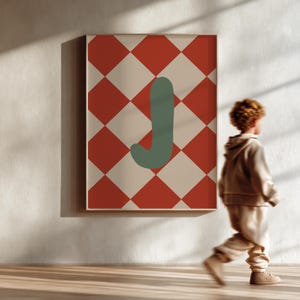 Op de afbeelding: Een ingelijste kunstprint met een rood en crèmekleurig ruitpatroon en een groene letter "J". Het kunstwerk wordt op een witte muur getoond. Een kind loopt voor het kunstwerk.
