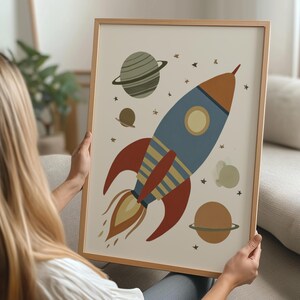 Peut inclure: Une affiche encadrée représentant une fusée de dessin animé dans l'espace. La fusée est bleue, orange et rouge, avec des flammes jaunes. Des planètes et des étoiles entourent la fusée sur un fond crème. Le cadre est de couleur bois clair.