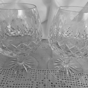 May include: Two clear crystal brandy snifters with diamond and floral cut patterns. The glasses have a rounded bowl shape and sit on a starburst base. The background is a white cloth and a lace tablecloth.