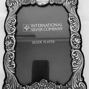 May include: A silver-plated picture frame with an ornate, embossed border featuring floral rose motifs and scrollwork. The center glass displays the text INTERNATIONAL SILVER COMPANY and SILVER PLATED in white lettering against a dark background.