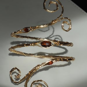 May include: Gold-toned arm cuff with a swirling design. The cuff features three bands with amber-colored gemstones. The ends of the cuff have spiral accents. The jewelry is displayed on a white surface.