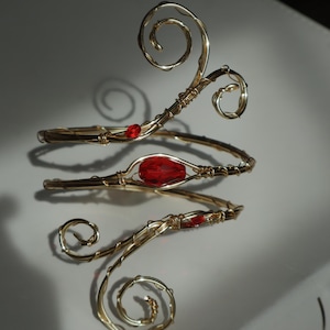 May include: Gold-toned arm cuff with a spiral design and red accents. The cuff features a central teardrop-shaped red stone and smaller red beads. The wire-wrapped design creates an elegant, artistic look, suitable for special occasions.