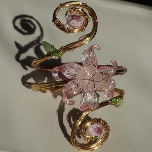 May include: A gold-coloured wire bracelet with a pink flower and two spiralling accents. The flower has delicate pink petals. Green leaves and pink orbs add detail. The bracelet is likely for decorative wear.