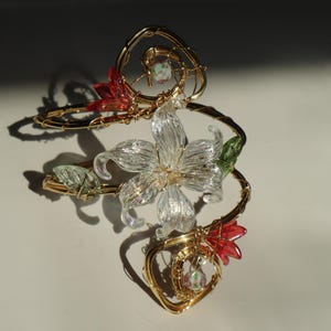 May include: A gold-toned wire floral arm cuff with a large, clear flower at the center. Red and green accents are placed around the cuff, with a heart-shaped design. The cuff is designed to be worn on the upper arm.
