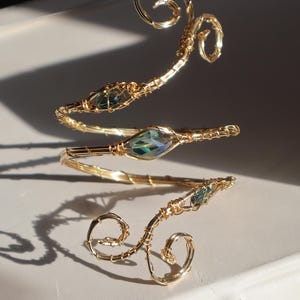 Puede incluir: Un brazalete dorado con un diseño en espiral, con tres piedras preciosas iridiscentes en forma de lágrima. El brazalete está hecho de alambre y descansa sobre una superficie blanca, mostrando su intrincada artesanía.