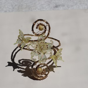 May include: A gold-coloured wirework bracelet with a floral design, iridescent petals, and butterfly accents. The bracelet has a spiral design with small, round, pearl-like embellishments. The overall aesthetic is delicate and nature-inspired.