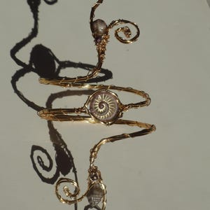 May include: A gold-toned wire arm cuff with a spiral design. The cuff features a central, circular, light-colored stone with a spiral pattern, and is adorned with small, clear and purple stones. The design includes decorative swirls.
