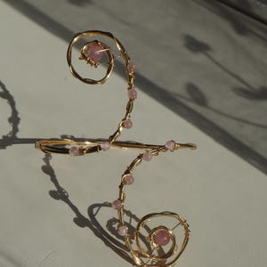 Op de afbeelding: Een goudkleurige draadarmmanchet met een spiraalontwerp en roze kralen. De manchet heeft een delicate, handgemaakte uitstraling, waarbij de kralen een vleugje kleur en textuur toevoegen. Het ontwerp is ingewikkeld en artistiek.