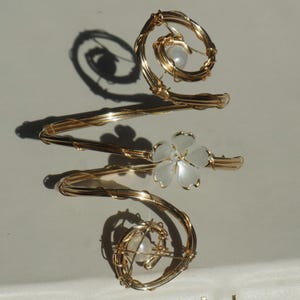 Puede incluir: Brazalete dorado con diseño en espiral, con una flor blanca y dos perlas. El brazalete se envuelve alrededor del brazo con una estructura delicada, similar a un alambre. El diseño es elegante y decorativo.