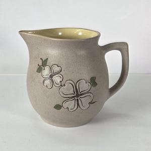 May include: A tan ceramic pitcher with a handle and a spout. The pitcher features a floral design in white and green. The interior of the pitcher is yellow. The pitcher is likely used for serving beverages.