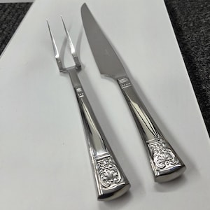 May include: A silver carving set, including a fork and a knife, with ornate floral detailing on the handles. The set is displayed on a white surface, showcasing the polished finish of the utensils.