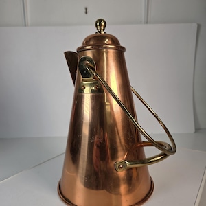May include: A polished copper coffee pot with a conical body, featuring a brass swing handle attached to the sides and a brass lid topped with a small spherical finial. The vessel has a wide base that tapers toward the top, showcasing a warm metallic finish with visible reflections on its smooth, curved surface.